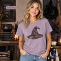 A woman wearing a dusty purple graphic t-shirt with a witch hat design, standing in a cozy, darkly decorated Halloween-themed room with candles, a cauldron, and broomsticks.