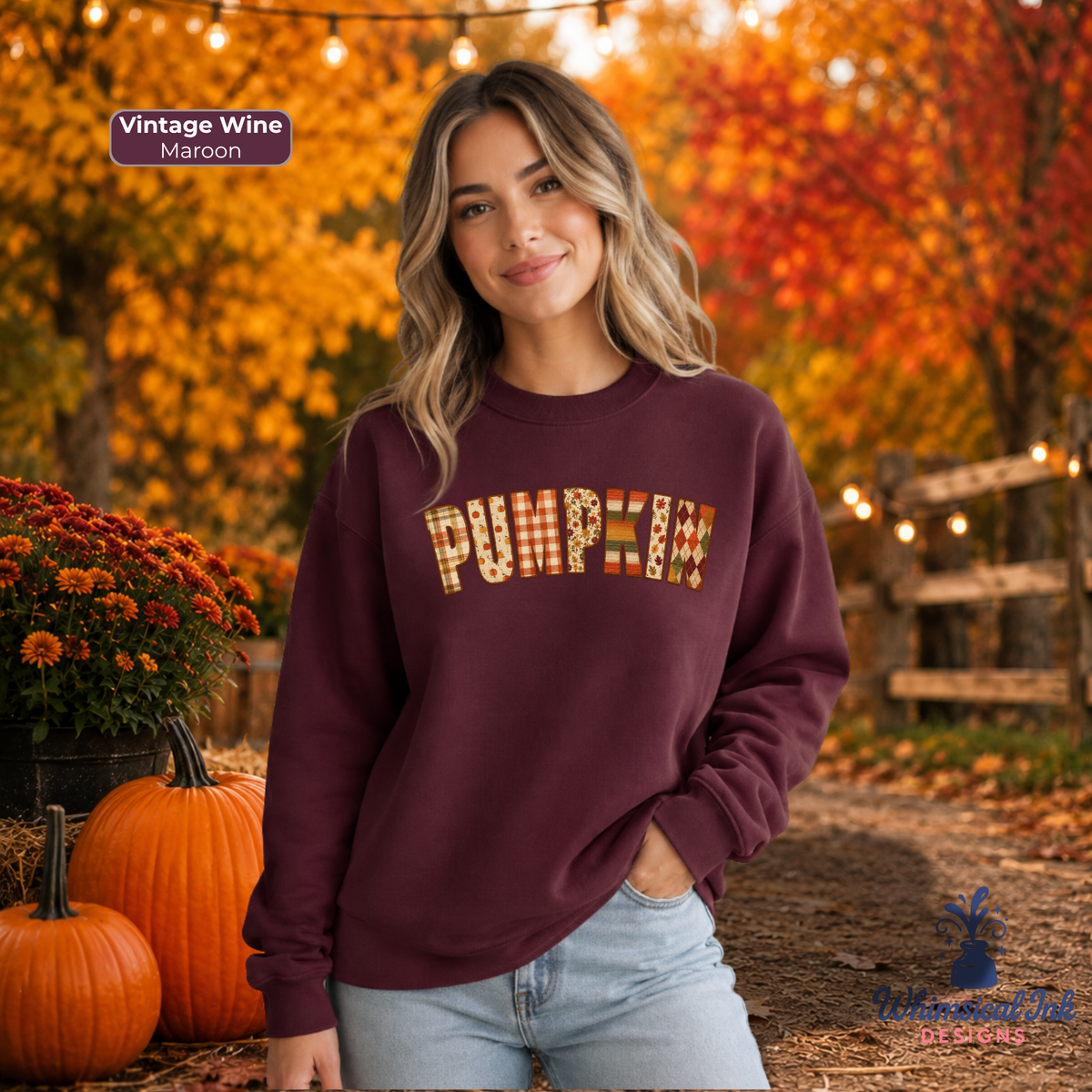 A woman wearing a maroon sweatshirt with a 'Pumpkin' text graphic made of autumn-themed patterns, standing outdoors in a fall garden with pumpkins and orange foliage.