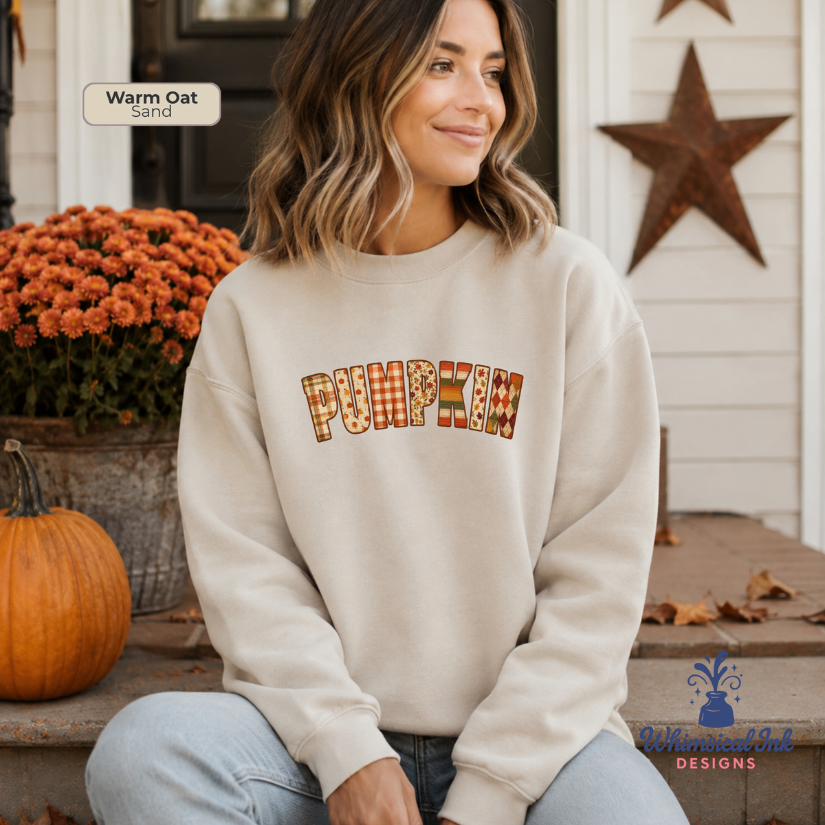 A woman wearing a cream-colored crewneck sweatshirt with 'PUMPKIN' written across the chest in a fall-themed patterned font. She is sitting on a porch with a pumpkin, orange flowers, and a wooden star in the background.