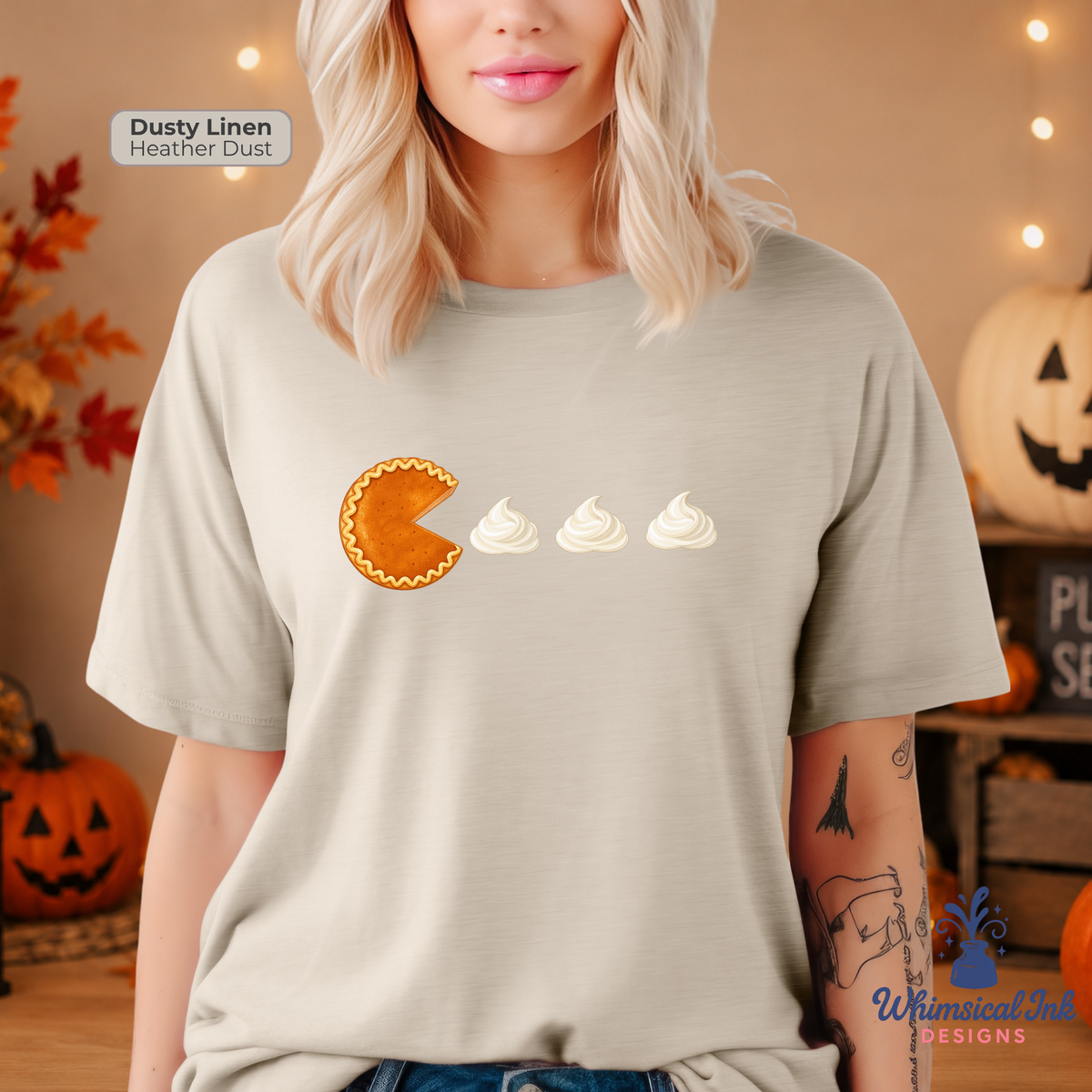 A woman wearing a light heather dust gray t-shirt with a graphic of a pumpkin pie eating three swirls of whipped cream. The image is set in a cozy, Halloween-themed room with pumpkins and string lights, and features a 'Dusty Linen Heather Dust' color label and a 'Whimsical Ink Designs' watermark.