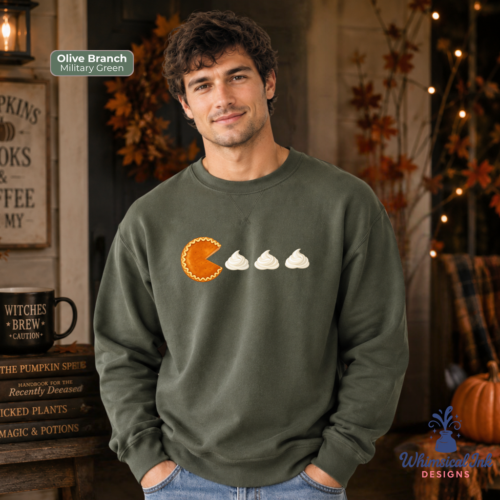 A man wearing an olive branch crewneck sweatshirt with a graphic of a pie and three whipped cream illustrations. The background is a cozy, autumn-themed room with fall decorations, a pumpkin, and a sign that reads 'Olive Branch Military Green'.