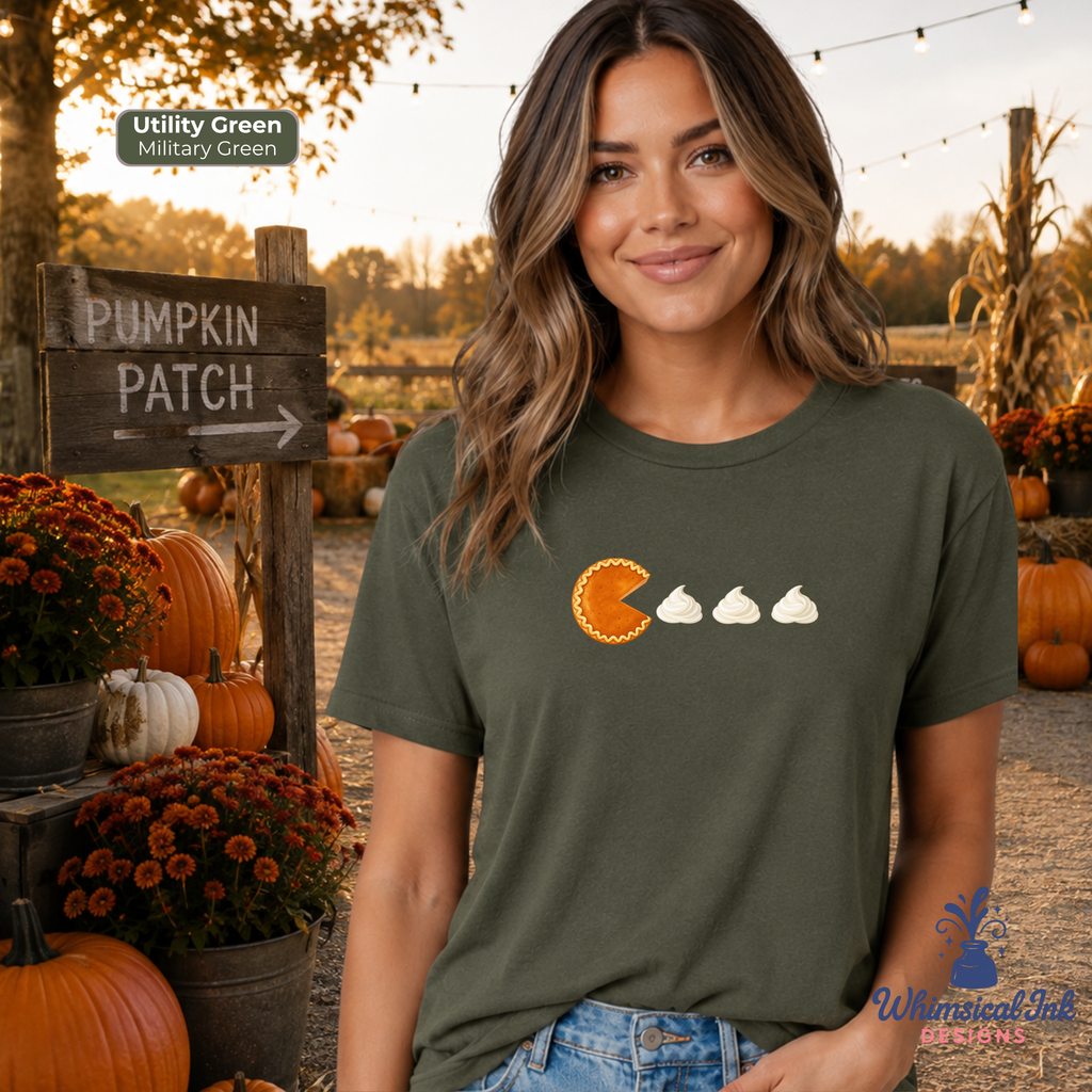 A woman wearing an olive green t-shirt with a graphic of a pie eating whipped cream, standing in a pumpkin patch at sunset. The image includes a color label indicating the shirt color is 'Utility Green' and a brand logo for 'Whimsical Vibes Designs'.