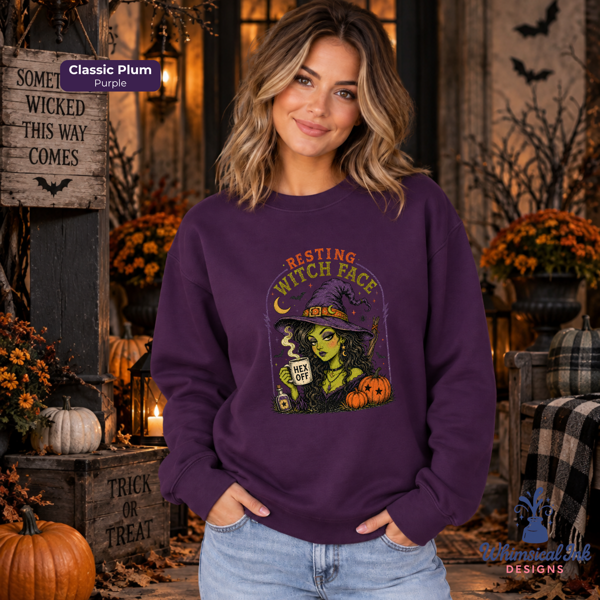A woman wearing a purple crewneck sweatshirt with a Halloween graphic of a witch holding a mug that says 'HEX OFF'. The background is a cozy, decorated autumn porch with pumpkins, candles, and fall foliage.