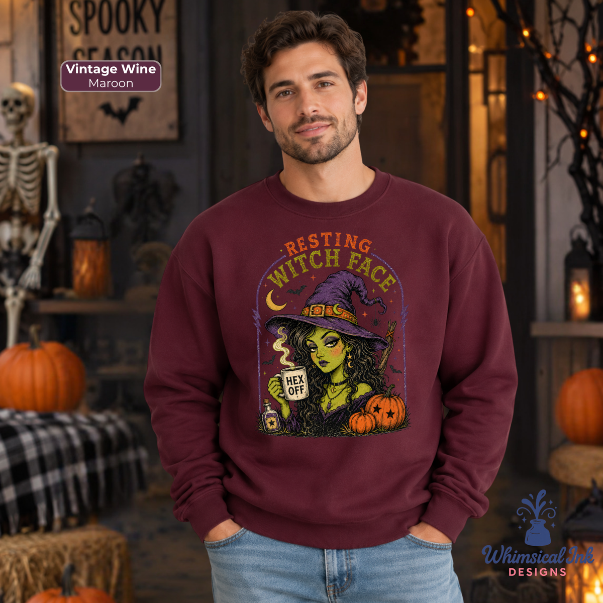 A man wearing a maroon crewneck sweatshirt with a colorful graphic of a witch holding a mug that says 'HEX OFF'. The background is a spooky Halloween-themed room with pumpkins, a skeleton, and warm lights.