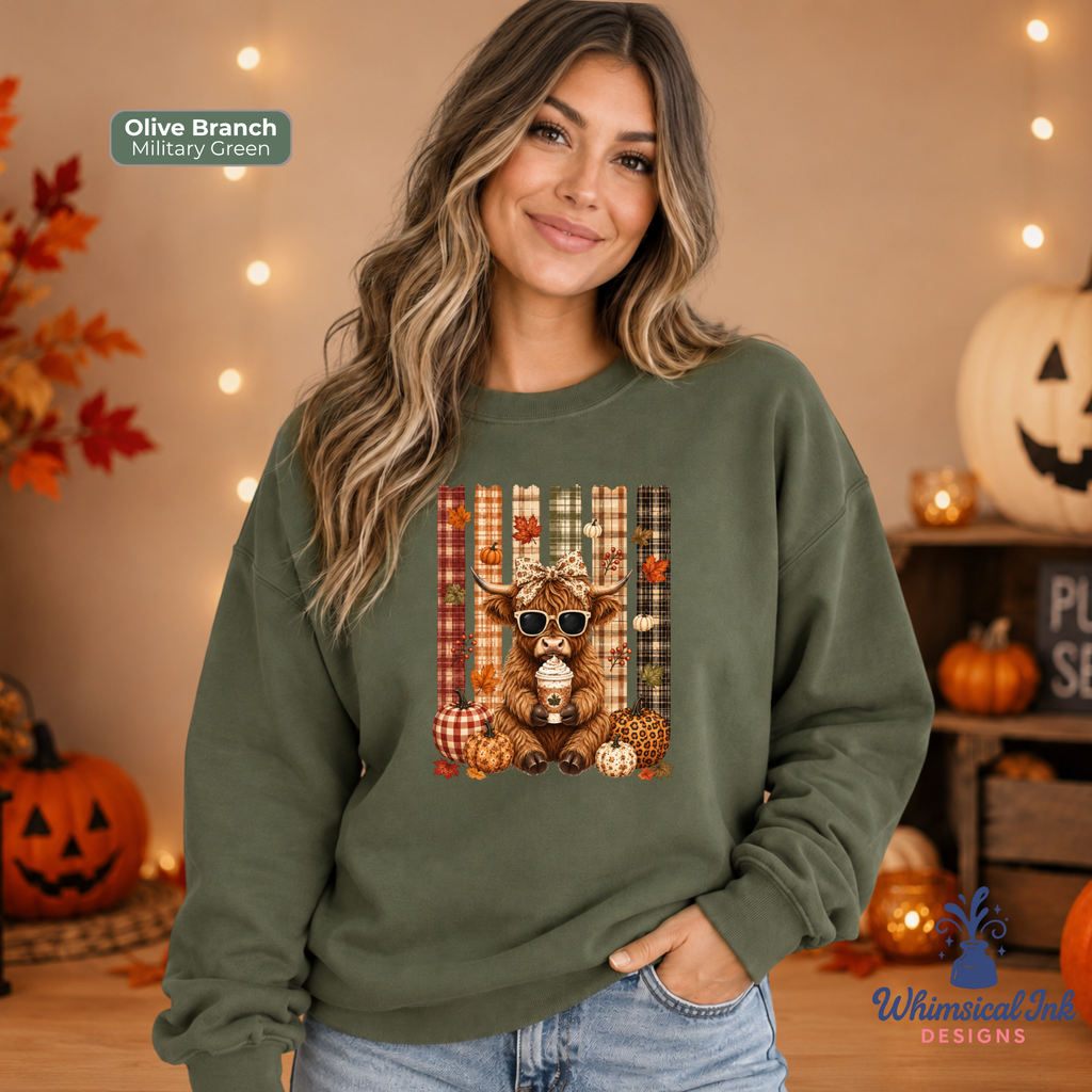A woman wearing an olive branch military green crewneck sweatshirt with a central graphic of a highland cow wearing sunglasses, surrounded by pumpkins and plaid patterns. The background is a cozy, warm autumn setting with string lights, pumpkins, and fall foliage.