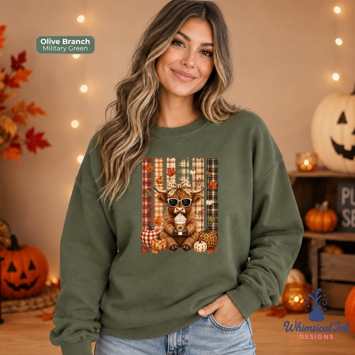 A woman wearing an olive branch military green crewneck sweatshirt with a central graphic of a highland cow wearing sunglasses, surrounded by pumpkins and plaid patterns. The background is a cozy, warm autumn setting with string lights, pumpkins, and fall foliage.