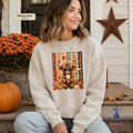 A woman wearing a cream-colored sweatshirt with a fall-themed graphic of a highland cow wearing sunglasses, sitting on a porch with pumpkins and orange flowers in the background.