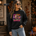 A woman modeling a black crewneck sweatshirt with a colorful graphic that reads 'SAVE A HORSE RIDE A BROOMSTICK' in purple, pink, and yellow. The photo is set in a cozy, autumn-themed rustic room with fall decorations, a pumpkin, and warm string lights.