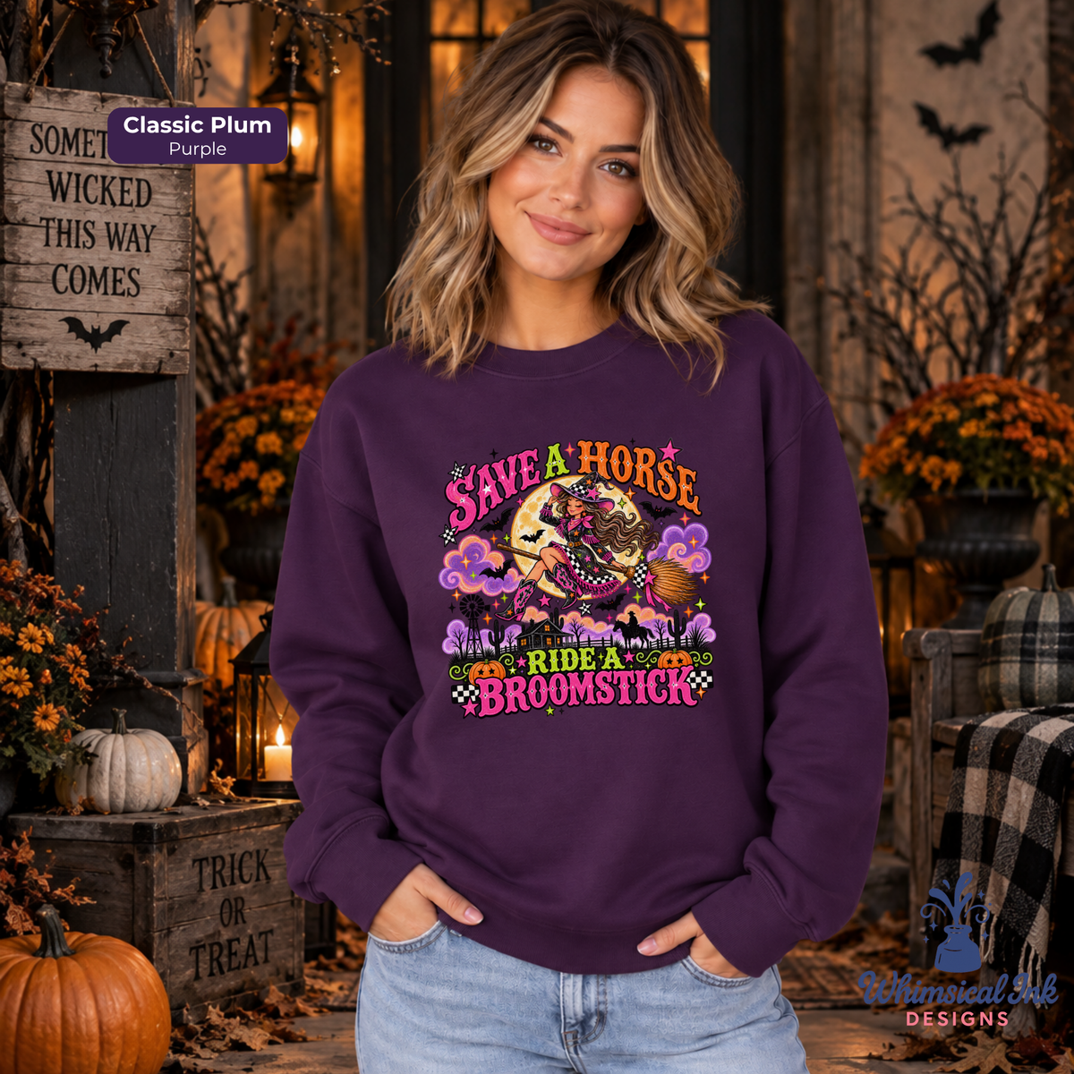 A woman wearing a plum purple graphic sweatshirt with a colorful 'Save a Horse Ride a Broomstick' Halloween design, posed in a cozy, decorated autumn-themed room.