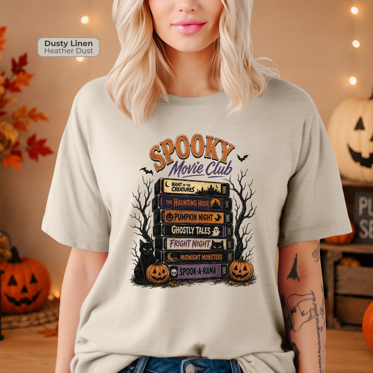 A woman wearing a light beige heather dust t-shirt with a 'Spooky Movie Club' graphic print, posed in a cozy Halloween-themed room with pumpkins and string lights.