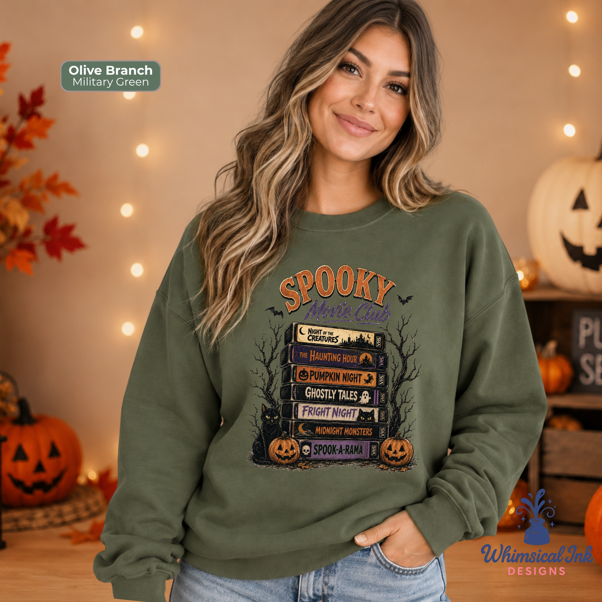 A woman wearing an olive branch green crewneck sweatshirt with a 'Spooky Night' graphic print, posed in a cozy, warmly lit room with pumpkins and string lights.