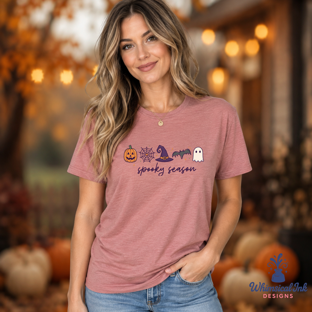 A woman wearing a heathered dusty rose short-sleeve t-shirt with a Halloween graphic that reads 'spooky season' and features pumpkins, a spiderweb, a witch hat, a bat, and a ghost. The background is a warm, blurred autumn porch scene with pumpkins.