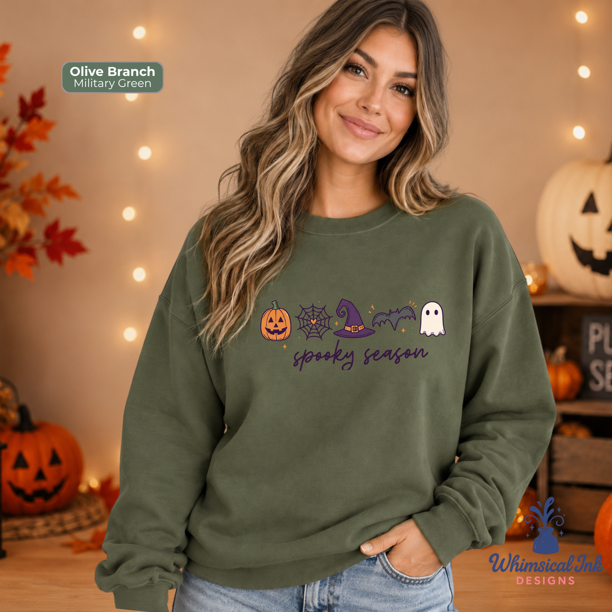 A woman wearing an olive branch military green sweatshirt with a 'spooky season' Halloween graphic, posed in a cozy, warmly lit fall-themed room with pumpkins and string lights.
