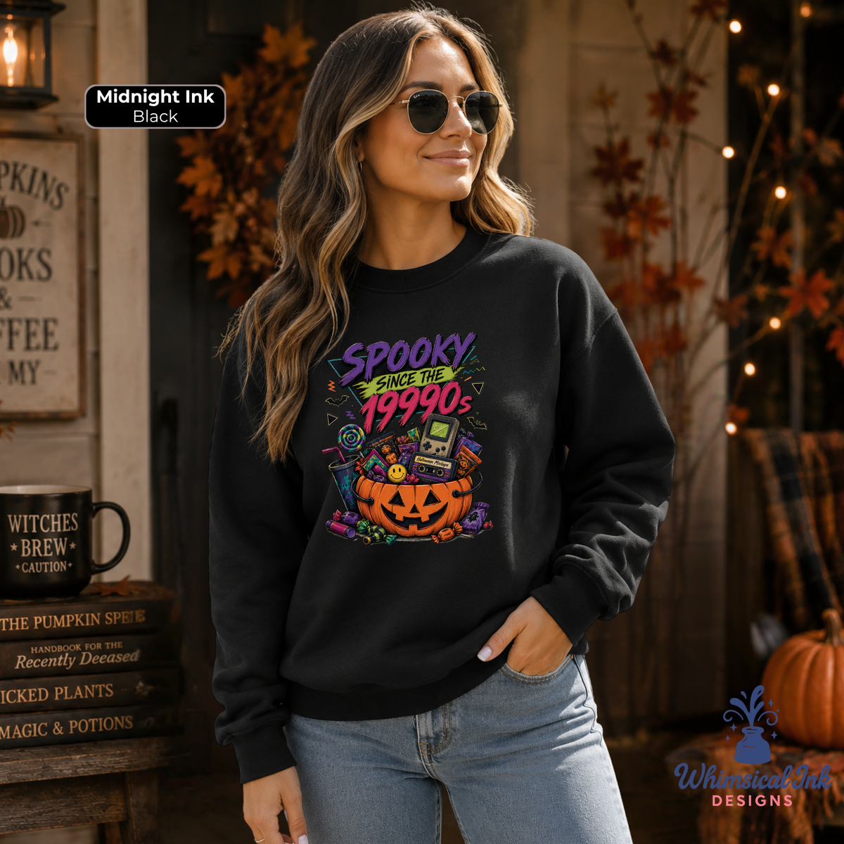 A woman wearing a black crewneck sweatshirt with a colorful 'Spooky Since the 1990s' graphic print featuring a jack-o'-lantern and retro computer. The image is set in a cozy, autumn-themed room with fall decorations, and includes a 'Midnight Ink Black' label overlay.