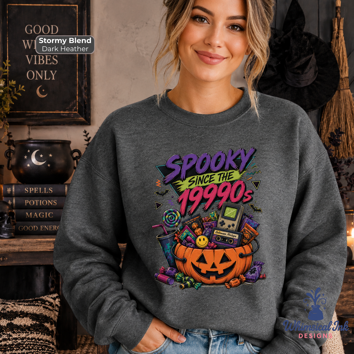 A woman wearing a dark gray crewneck sweatshirt with a colorful 'Spooky Since the 1990s' graphic print featuring a jack-o'-lantern, candy corn, and a retro game console. The background is a cozy, dark-themed Halloween room with a cauldron, broom, and witch hat.