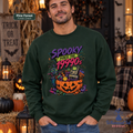 A man wearing a dark forest green crewneck sweatshirt with a colorful graphic that reads 'Spooky Since The 1990s' above a jack-o'-lantern and retro game console. The background is a cozy, warmly lit Halloween-themed room with pumpkins, lanterns, and a 'Trick or Treat' sign.