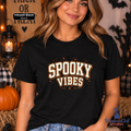 A woman wearing a black graphic t-shirt with 'SPOOKY VIBES' printed in a distressed cream and orange font, set against a Halloween-themed background with pumpkins and lanterns.