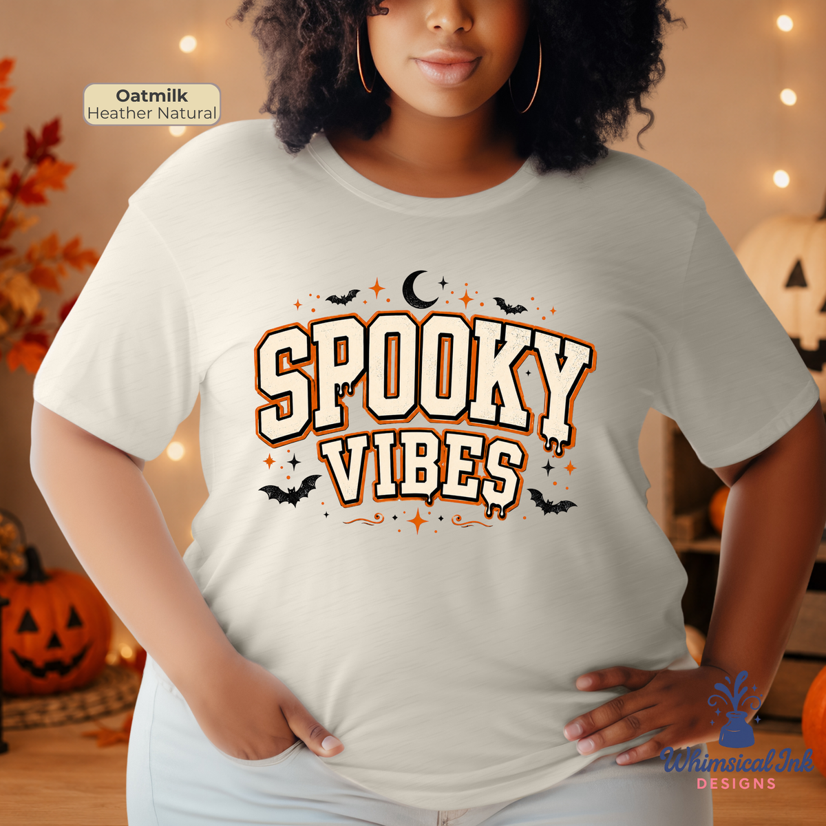 A woman wearing a light oatmeal-colored graphic t-shirt with 'Spooky Vibes' printed in a stylized font with bats, a crescent moon, and stars. The background is a cozy, warm Halloween-themed room with pumpkins and string lights.