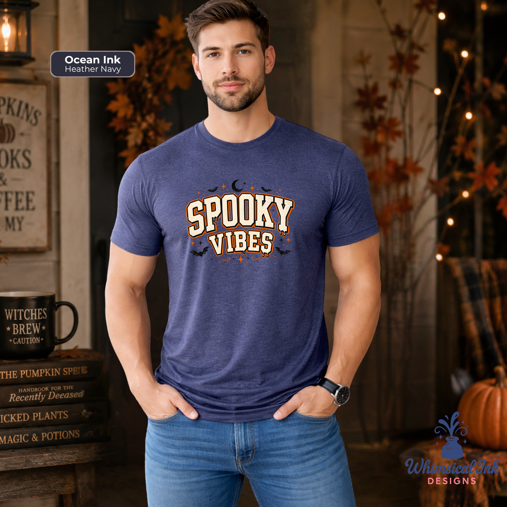 A man wearing a heather navy blue graphic t-shirt with 'SPOOKY VIBES' printed in cream and orange with black bats, set against a cozy, rustic autumn-themed background. A badge in the top left reads 'Ocean Ink Heather Navy'.