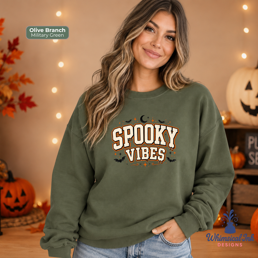 A woman wearing an olive green crewneck sweatshirt with a 'Spooky Vibes' graphic, posed in a cozy, warmly lit Halloween-themed room with pumpkins and string lights.