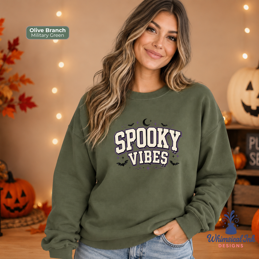 A woman wearing an olive green crewneck sweatshirt with 'SPOOKY VIBES' printed in white and black on the front, posed in a cozy, warmly lit Halloween-themed room with pumpkins and string lights.