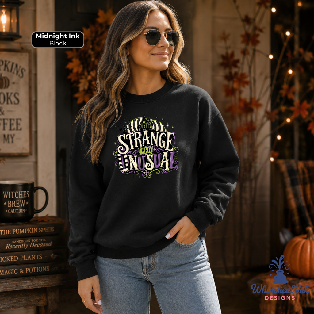 A woman wearing a black crewneck sweatshirt with a colorful graphic that reads 'STRANGE AND UNUSUAL'. She is wearing blue jeans and sunglasses, standing in a cozy, autumn-themed setting with pumpkins, fall leaves, and string lights.