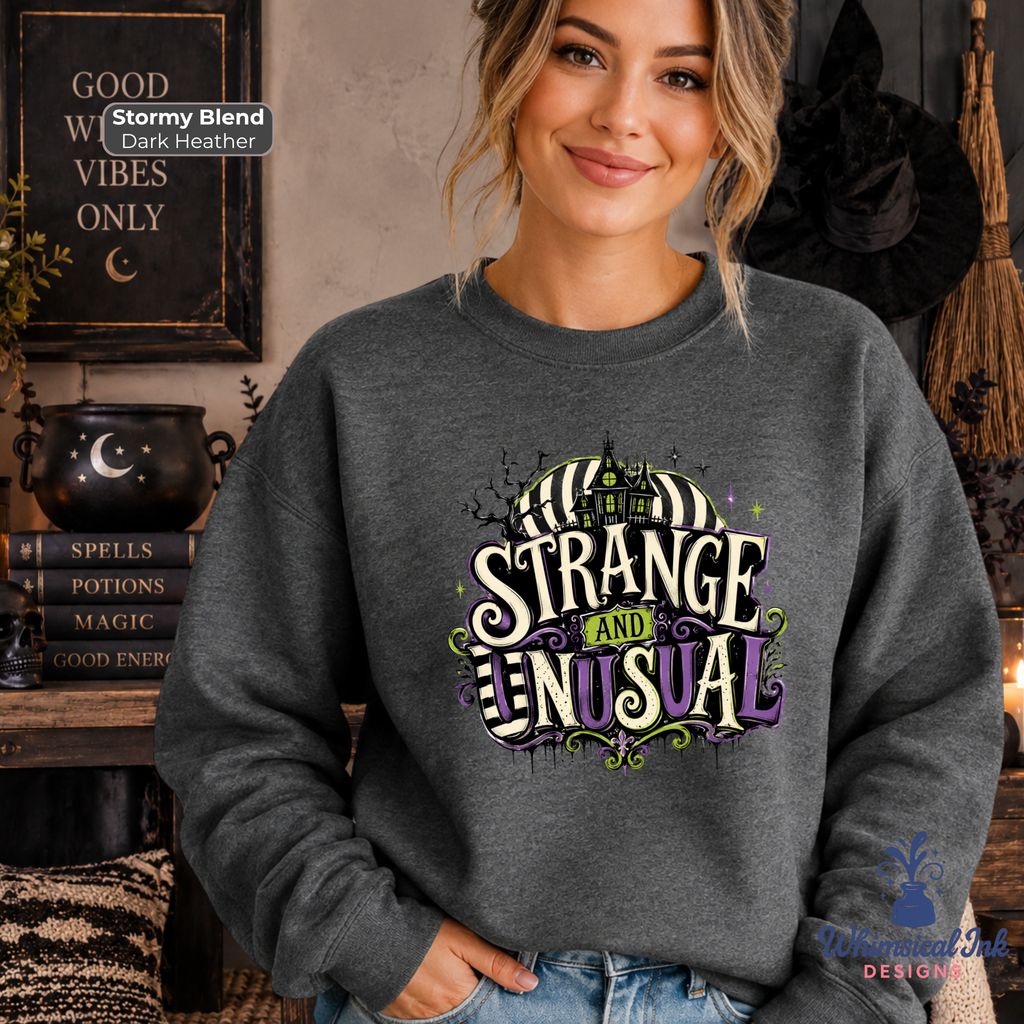 A woman wearing a dark heather gray crewneck sweatshirt with a colorful graphic that reads 'IT'S STRANGE AND UNUSUAL' in a spooky Halloween font. She is standing in a cozy, themed room with witch hats, a cauldron, and books.