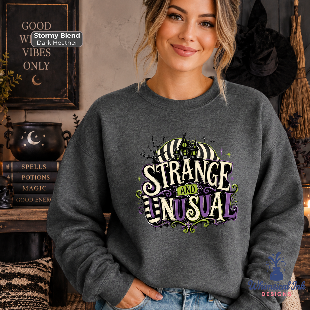 A woman wearing a dark heather gray crewneck sweatshirt with a colorful graphic that reads 'IT'S STRANGE AND UNUSUAL' in a spooky Halloween font. She is standing in a cozy, themed room with witch hats, a cauldron, and books.