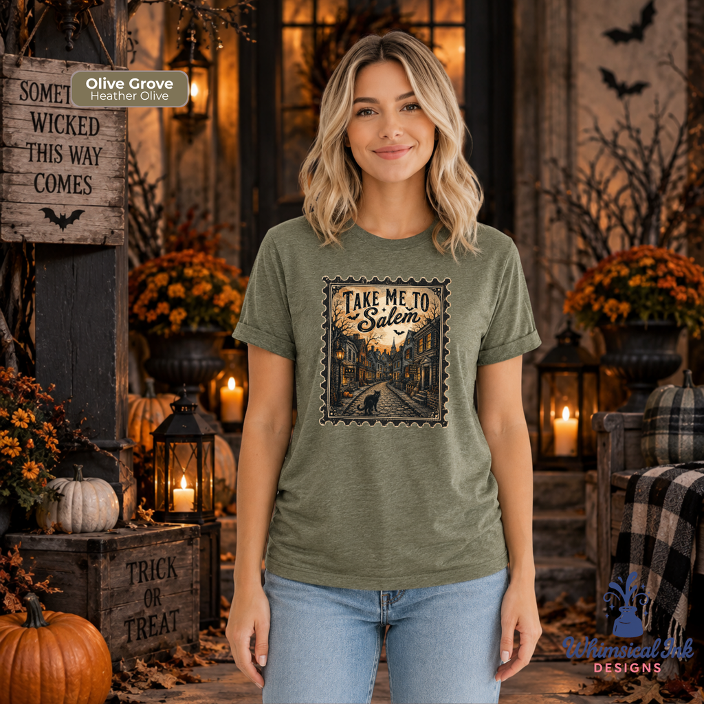 A woman wearing an olive green graphic t-shirt with a vintage-style 'Take Me To Salem' illustration, standing in a cozy, decorated autumn-themed room with pumpkins, candles, and fall foliage. An 'Olive Grove Heather Olive' label is visible in the top left corner.