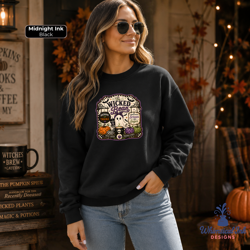 A woman wearing a black crewneck sweatshirt with a 'Wickedly Cute' Halloween graphic, standing in a cozy autumn-themed room. The sweatshirt features a colorful graphic of a ghost, pumpkin, witch hat, and cauldron under a coffee shop sign that reads 'The Wicked Bean'.