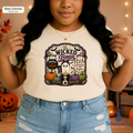 A woman wearing a cream-colored graphic t-shirt with a colorful 'The Wicked Bean' Halloween design, paired with light-wash distressed denim jeans. The background is a cozy, warm-toned indoor setting with string lights and pumpkins.