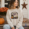 A woman sits on porch steps wearing a cream sweatshirt with a colorful Halloween graphic that reads 'Wicked Beautiful.' The scene includes a pumpkin, orange flowers, a rustic door, and a star decoration.