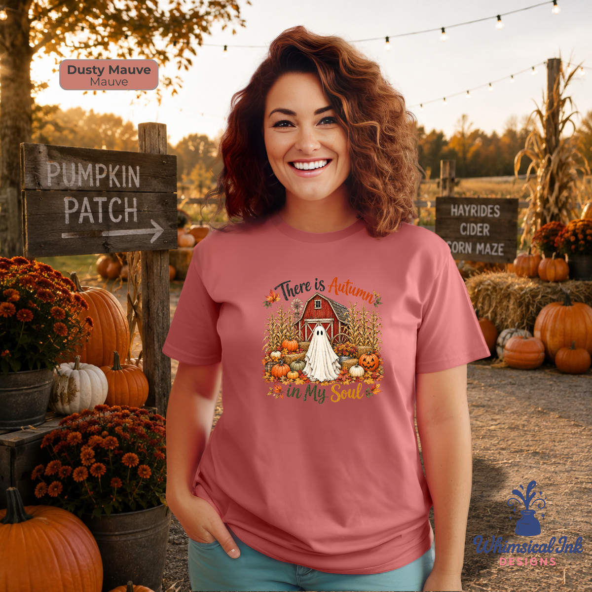 A woman stands outdoors at a fall-themed gathering wearing a dusty mauve graphic t-shirt with a fall harvest design. The shirt features a red barn, white ghost, pumpkins, and the text 'There is Autumn in My Soul'.