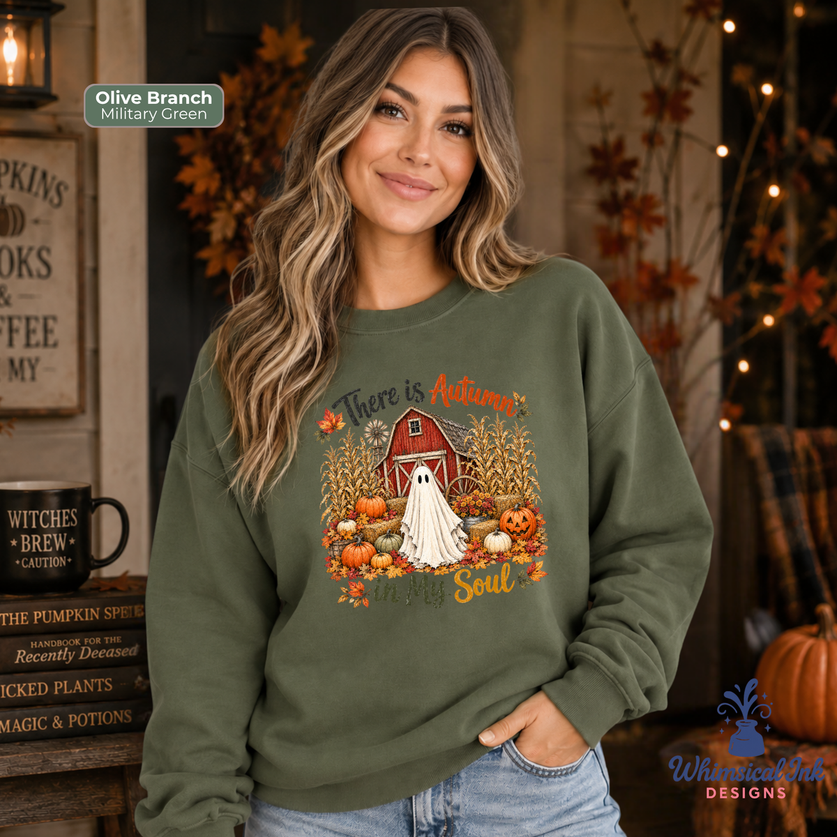 A woman wearing an olive branch military green sweatshirt with a fall-themed graphic that reads 'There is Autumn in My Soul' featuring a ghost in a white gown in front of a red barn surrounded by pumpkins and cornstalks. The background depicts a cozy autumn-themed room with warm lights and fall decor.