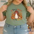 A woman wearing a sage green graphic t-shirt with a Halloween autumn design featuring a ghost, barn, pumpkins, and the text 'There is Autumn in My Soul'.