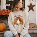 A woman wearing a cream-colored sweatshirt with a fall-themed graphic that reads 'There is Autumn in My Soul' featuring a ghost, barn, pumpkins, and cornstalks. She is sitting on a porch with pumpkins and orange flowers in the background.
