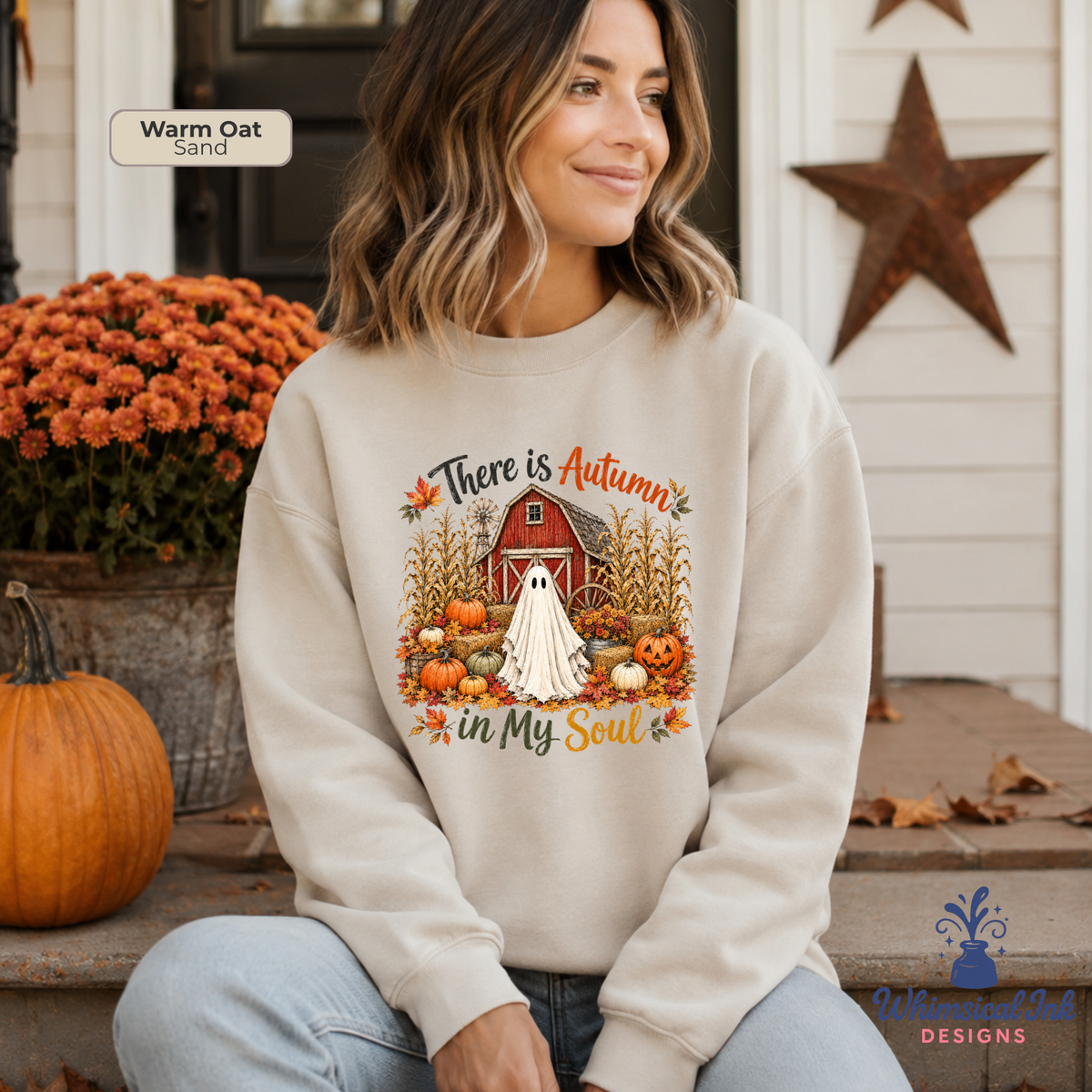 A woman wearing a cream-colored sweatshirt with a fall-themed graphic that reads 'There is Autumn in My Soul' featuring a ghost, barn, pumpkins, and cornstalks. She is sitting on a porch with pumpkins and orange flowers in the background.