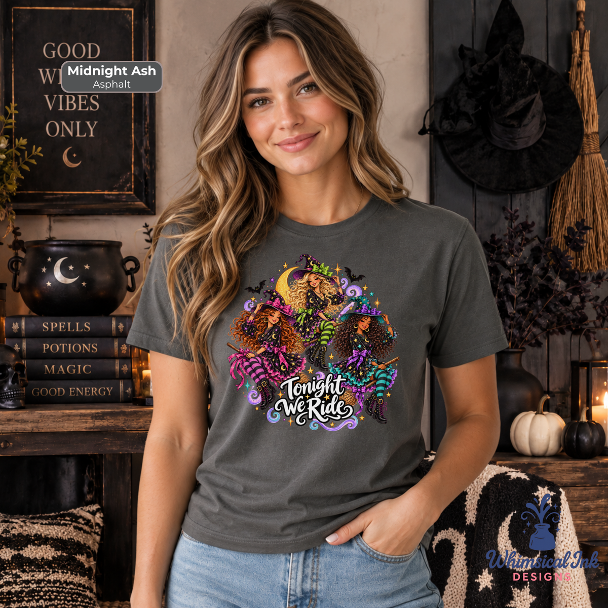 A woman wearing a dark gray graphic t-shirt with a colorful witch illustration and the text 'Tonight We Ride'. The background is a cozy, decorated Halloween-themed room with a black cauldron, books, and a broom.