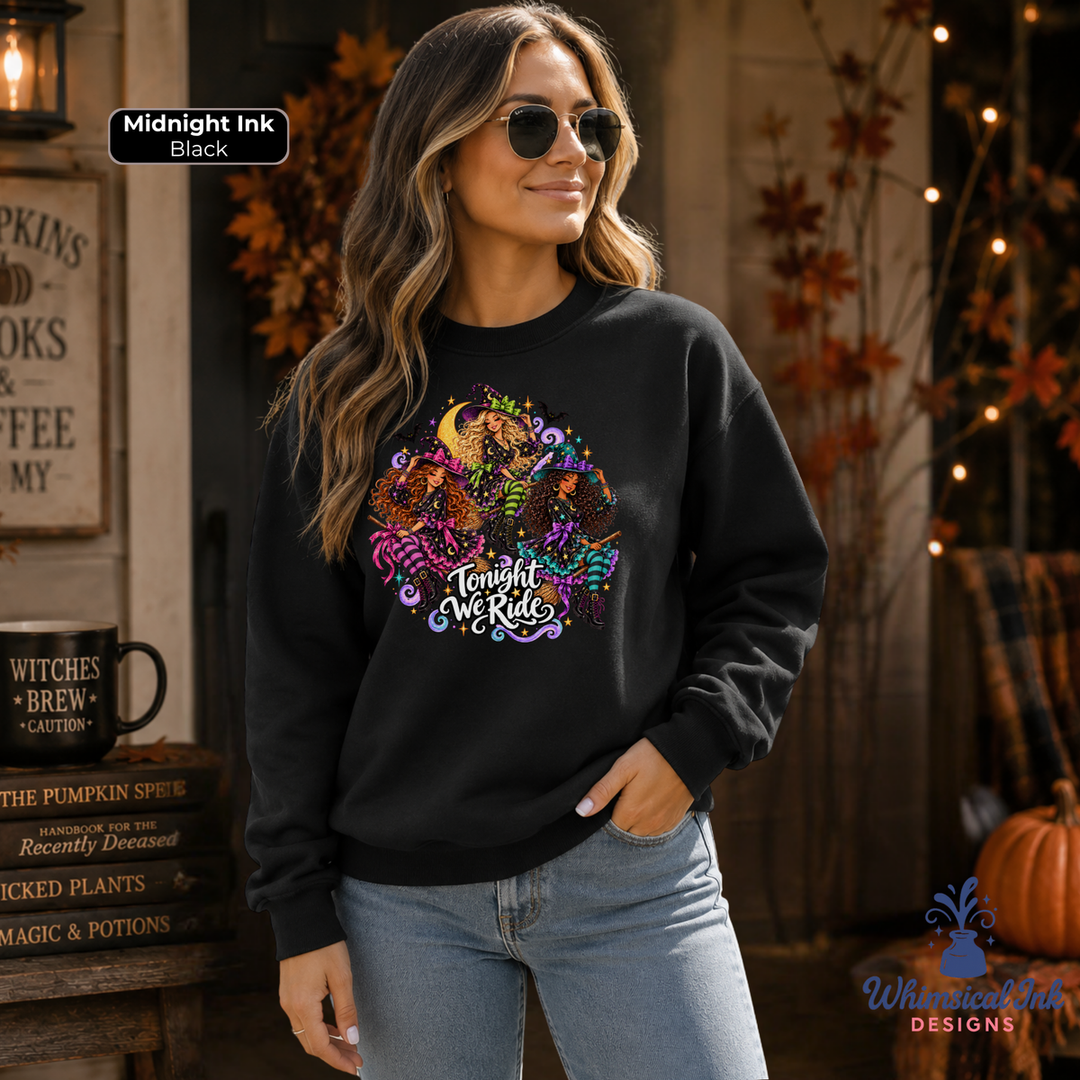 A woman wearing a black crewneck sweatshirt with a colorful graphic of pumpkins, a crescent moon, and the text 'Tonight We Ride' in white script. The background is a cozy, autumn-themed rustic shop setting with fall decorations, a pumpkin, and a sign that reads 'Midnight Ink Black'.