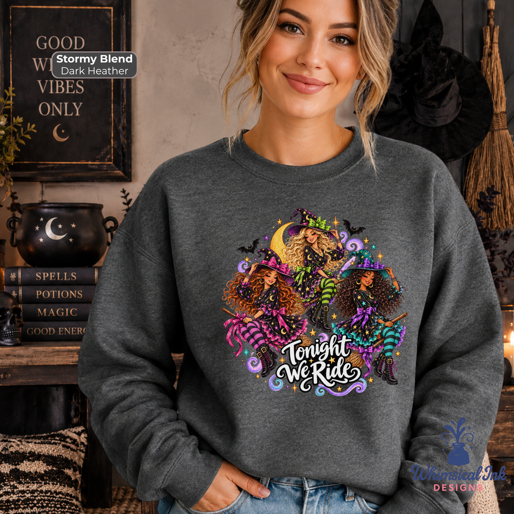 A woman wearing a dark heather gray crewneck sweatshirt with a colorful graphic of three witches and the text 'Tonight We Ride'. The background is a cozy, dark-themed room with Halloween decorations, including a black cauldron, witch hat, and broom.