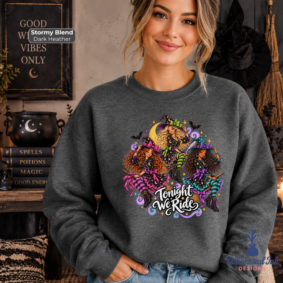 A woman wearing a dark heather gray crewneck sweatshirt with a colorful graphic of three witches and the text 'Tonight We Ride'. The background is a cozy, dark-themed room with Halloween decorations, including a black cauldron, witch hat, and broom.