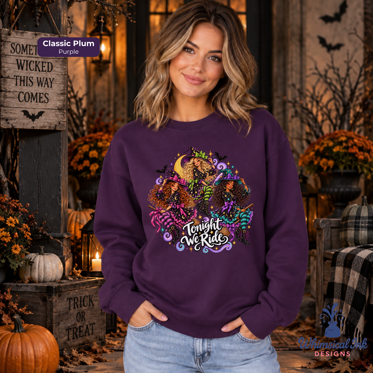 A woman wearing a purple crewneck sweatshirt with a colorful Halloween graphic that reads 'Tonight We Ride' with a three witches design. The background is a cozy, decorated autumn porch with pumpkins, candles, and fall foliage.