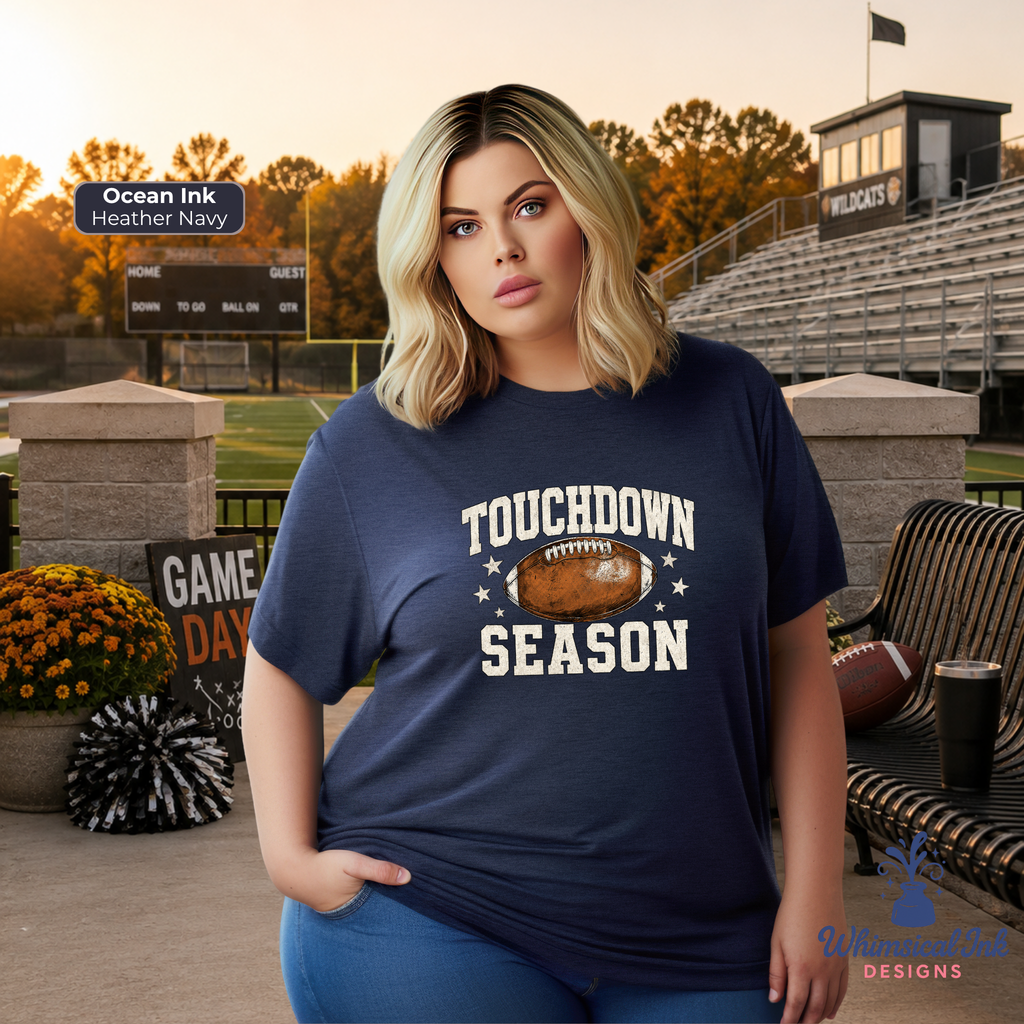A woman stands on a stadium set wearing a navy blue graphic T-shirt that reads 'TOUCHDOWN SEASON' with a football graphic in the center. The background includes bleachers, a scoreboard, flowers, and a bench.