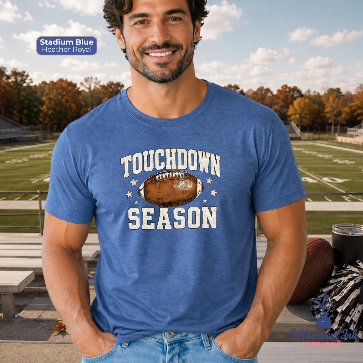 A man wearing a heather blue graphic t-shirt with 'TOUCHDOWN SEASON' and a football graphic printed on the front, standing on an outdoor football field. A blue label in the top left reads 'Stadium Blue Heather Royal'.
